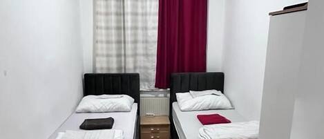 Una habitación pequeña y estrecha con dos camas individuales, piso de madera, una puerta blanca y una ventana con cortinas.