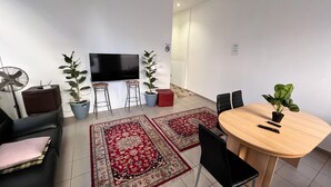 A modern living room with a flat-screen TV, two potted plants, a red ottoman, a dining table with chairs, and a large red and beige rug. - Hostel Wieden (Vienna)