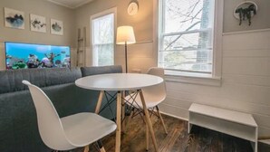 Una sala de estar con un sofá gris, una mesa redonda blanca y dos sillas blancas. Hay un televisor de pantalla plana en la pared, una lámpara de pie y una ventana con persianas.