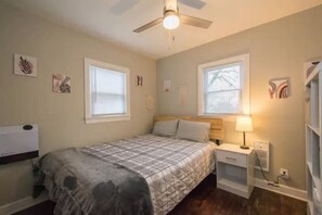 A bedroom with a bed, two windows, a ceiling fan, a bedside table with a lamp, and wall art.
