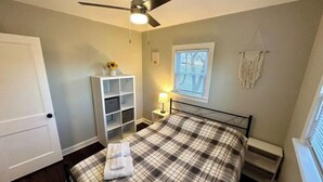 A bedroom with a bed, a ceiling fan, a window with blinds, a nightstand with a lamp, and a white shelving unit. - The Red Door Cottage (Hendersonville)
