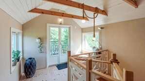 Une pièce spacieuse avec un plafond haut, des poutres en bois, un escalier et une grande fenêtre donnant sur un paysage verdoyant.