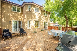 Une terrasse en bois avec des meubles d’extérieur, un barbecue et une maison en briques dotée de grandes fenêtres.