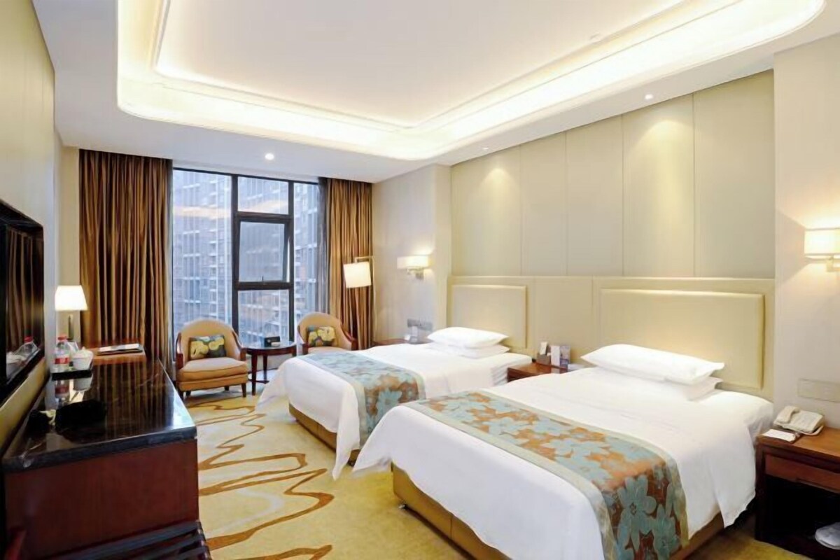 A hotel room with two beds, a desk, a chair, a TV, and a large window with curtains.