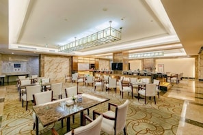 A spacious hotel lobby with multiple dining tables and chairs, a large seating area, and a high ceiling with a glass ceiling section.