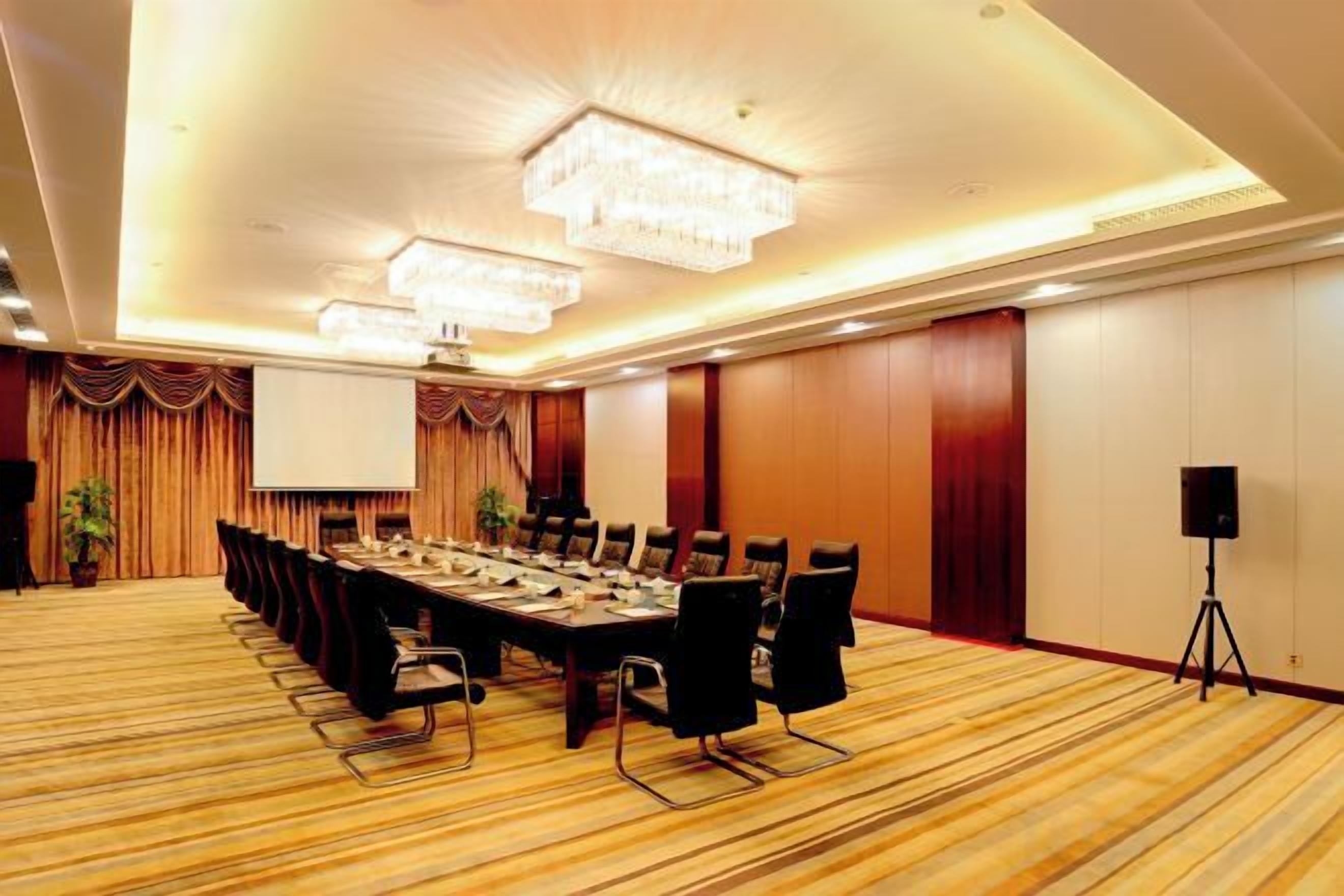 Un salón de conferencias con una larga mesa, sillas, una pantalla de proyector y un altavoz sobre un trípode.