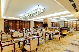 Amplia zona de comedor de hotel con varias mesas y sillas, una barra central y un techo alto con una lámpara de diseño geométrico.