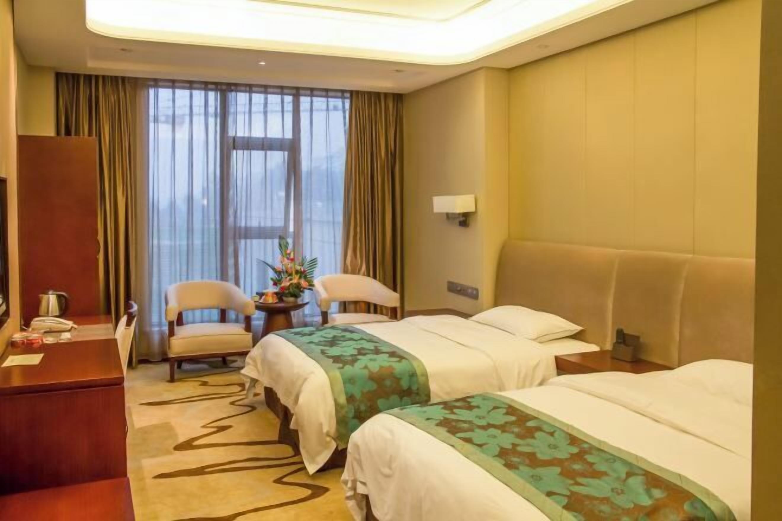Ein Hotelzimmer mit zwei Betten, einem Schreibtisch, einem Stuhl, einem kleinen Tisch mit einer Vase und einem Fenster mit Vorhängen.
