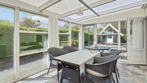 Un invernadero con mesa y sillas, ventanas grandes y vista a una casa.