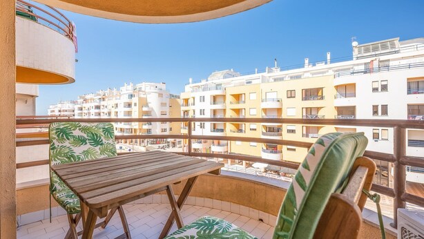 A balcony with a wooden table and chairs, a view of surrounding apartment buildings, and a clear blue sky. - GuestReady - Sunshine at Costa da Caparica (Costa da Caparica)