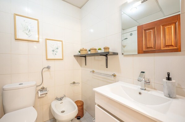 A bathroom with a toilet, bidet, vanity with a sink, wooden cabinets, and two framed pictures on the wall.