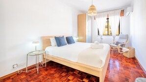 A modern bedroom with a wooden bed, a bedside table, a hanging light fixture, a chair, and a window with curtains. - GuestReady - Sunshine at Costa da Caparica (Costa da Caparica)
