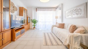 A modern living room with a flat-screen TV, wooden cabinets, a sofa, a potted plant, and a large window with sheer curtains. - GuestReady - Sunshine at Costa da Caparica (Costa da Caparica)