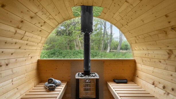 A wooden sauna with a central chimney, benches, and a view of a forest through an arched window. - Lovely home in Ulfborg with sauna (Ulfborg)