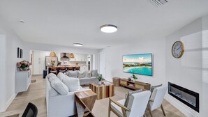 Ein modernes Wohnzimmer mit einer Couch, einem Couchtisch und einem Fernseher. Im Hintergrund befindet sich eine Küche mit Schränken und einem Essbereich.