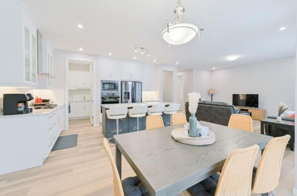 A modern kitchen with white cabinets, a dining area with a table and chairs, and a living area with a sofa and TV.