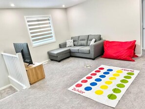 A living room with a grey sectional sofa, a red pillow, a white radiator, and a Twister mat on the floor.