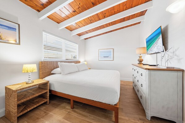 A bedroom with a wooden ceiling, a bed with white bedding, a bedside table with a lamp, a dresser, and a TV on the wall.