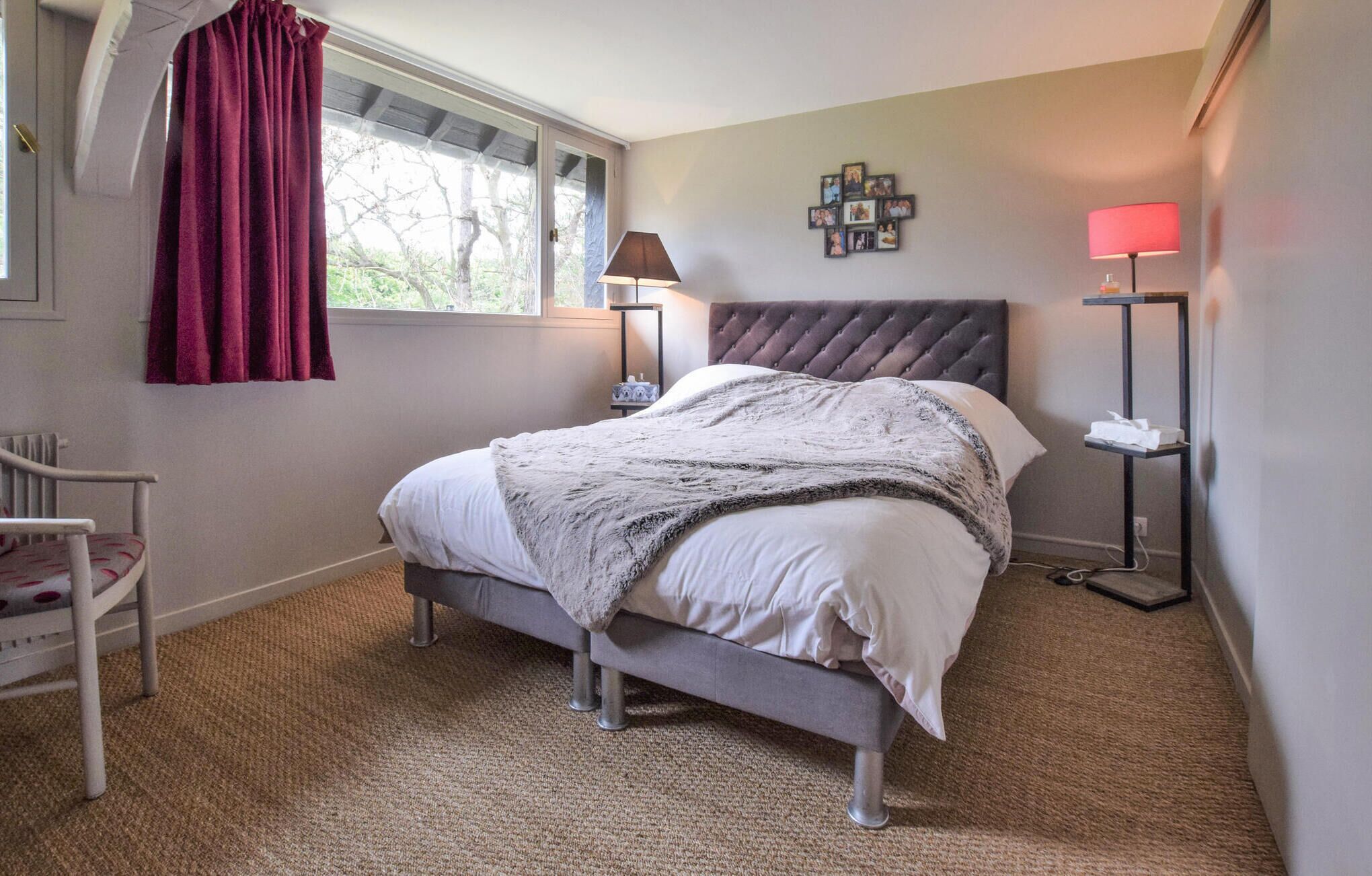 Une chambre à coucher avec un lit, une chaise, une fenêtre avec des rideaux, une lampe et un mur décoré de tableaux.