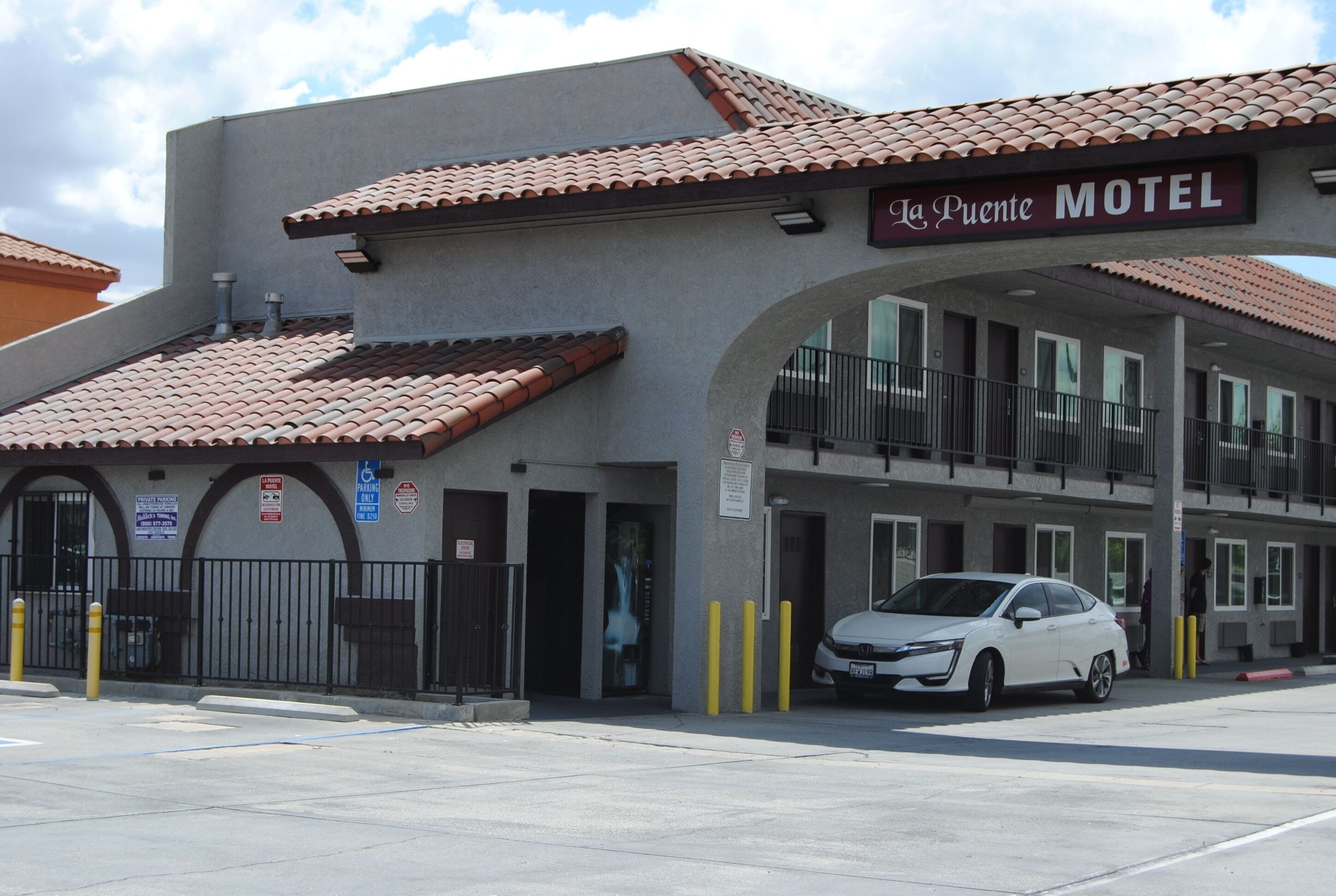 Un motel llamado 'La Puente' con techo de tejas, entrada protegida por una reja y un coche blanco estacionado al frente.