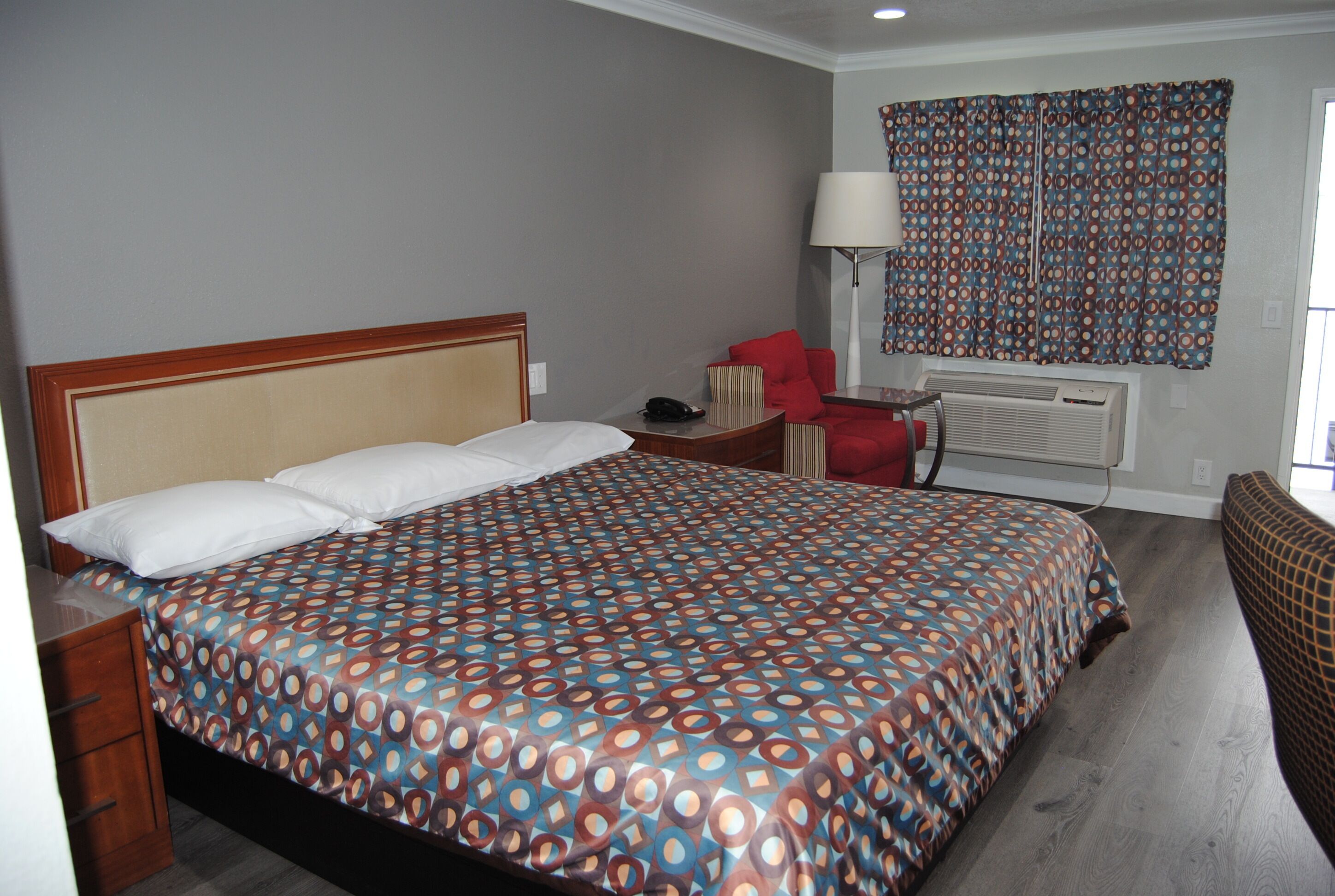 Habitación de hotel con una cama grande, un escritorio, una silla, una lámpara y una ventana con cortinas estampadas.