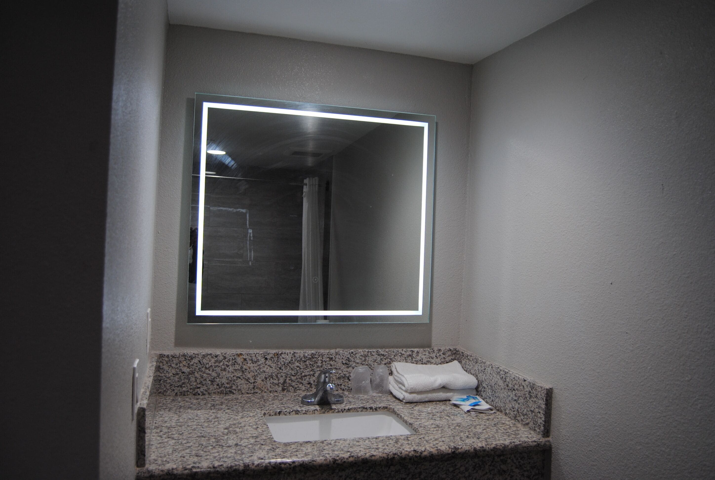 A bathroom with a granite countertop, a sink, a mirror with a light, and a towel on the counter.