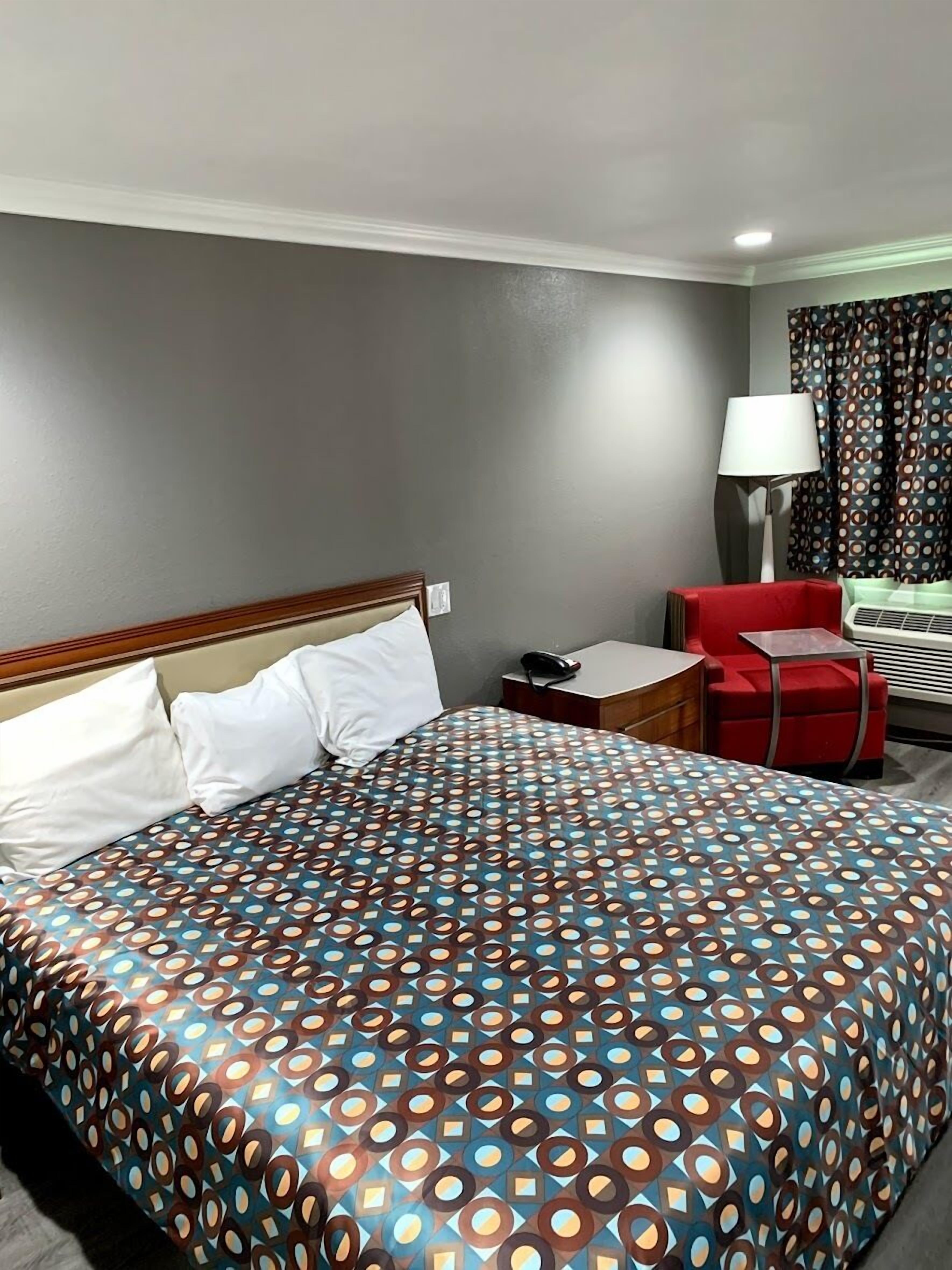 A hotel room with a bed, a red armchair, a small table with a lamp, and a window with curtains.