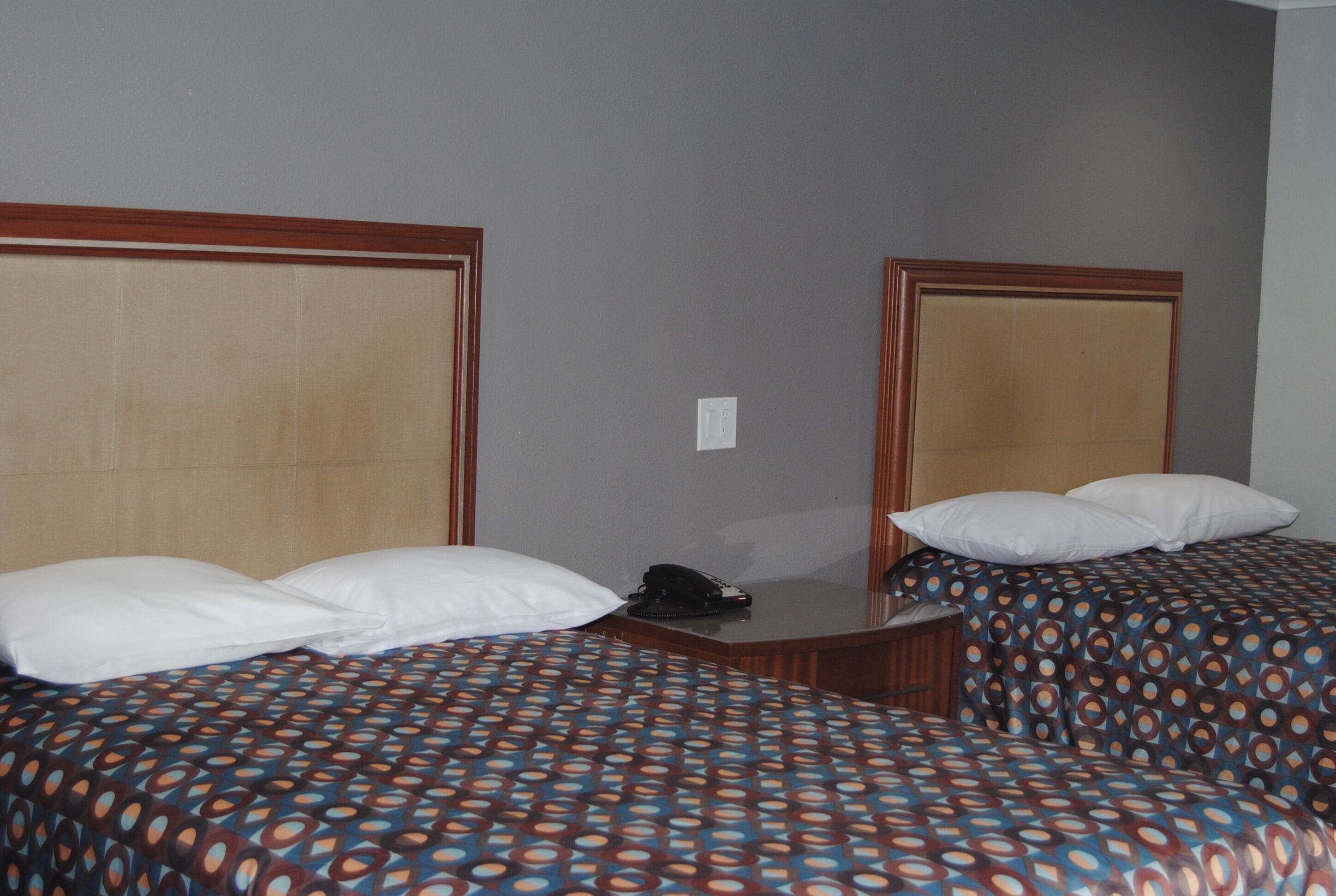 Habitación de hotel con dos camas, cabecero de madera, mesita de noche con teléfono y un enchufe en la pared.