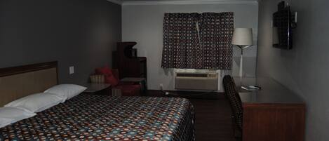 A hotel room with a bed, a desk, a chair, a lamp, and an air conditioning unit.