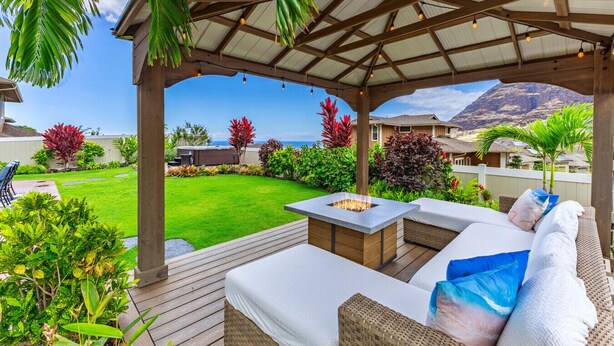 A covered outdoor seating area with a fire pit, surrounded by lush greenery and a view of the ocean and mountains. - Ocean Views|Beach 6 Min|Private Hot Tub|Gated|BBQ (Waianae)