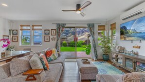 A spacious living room with a large sofa, a coffee table, and a television. There are multiple windows offering a view of a lush garden and mountains. - Ocean Views|Beach 6 Min|Private Hot Tub|Gated|BBQ (Waianae)