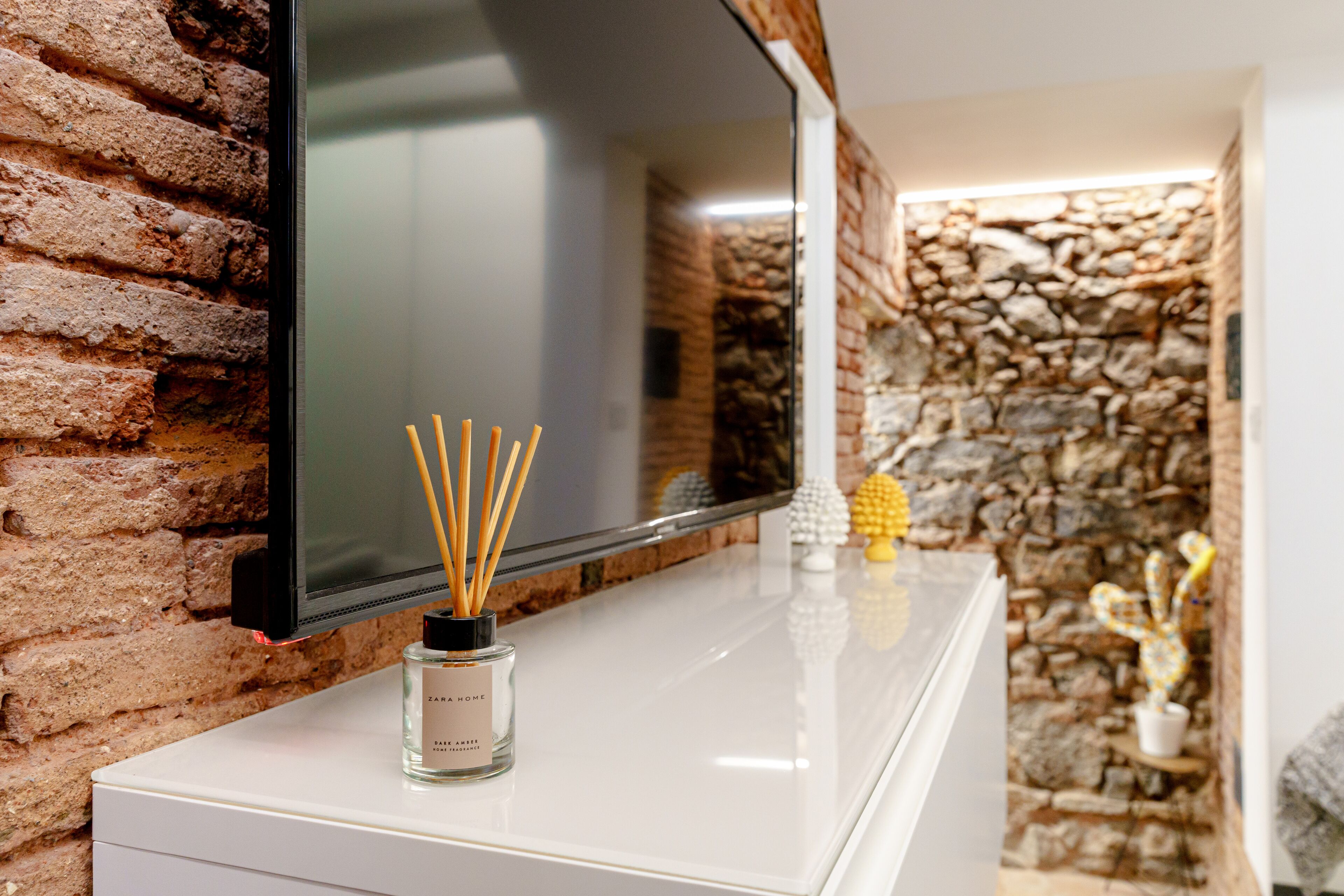 A flat-screen TV mounted on a brick wall, a white cabinet below, and a small glass bottle with reed sticks on top.