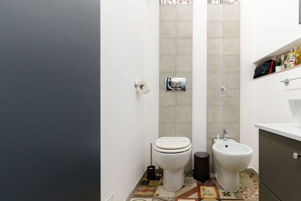 A bathroom with a toilet, bidet, and sink. - Lava rock formation in the center (Catania)