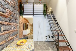 A room with a stone wall, a table with a yellow bowl, a staircase with a metal railing, and a view of a stone building. - Lava rock formation in the center (Catania)