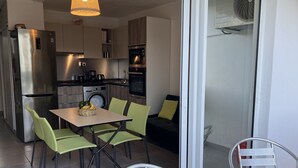 Une cuisine compacte dotée d’appareils électroménagers modernes, un coin repas avec des chaises vertes et une porte-fenêtre coulissante donnant sur un balcon.