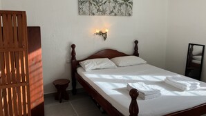 Un lit bien fait, avec du linge de lit blanc et une petite table de chevet, un climatiseur fixé au mur et un tableau aux motifs tropicaux au-dessus du lit.