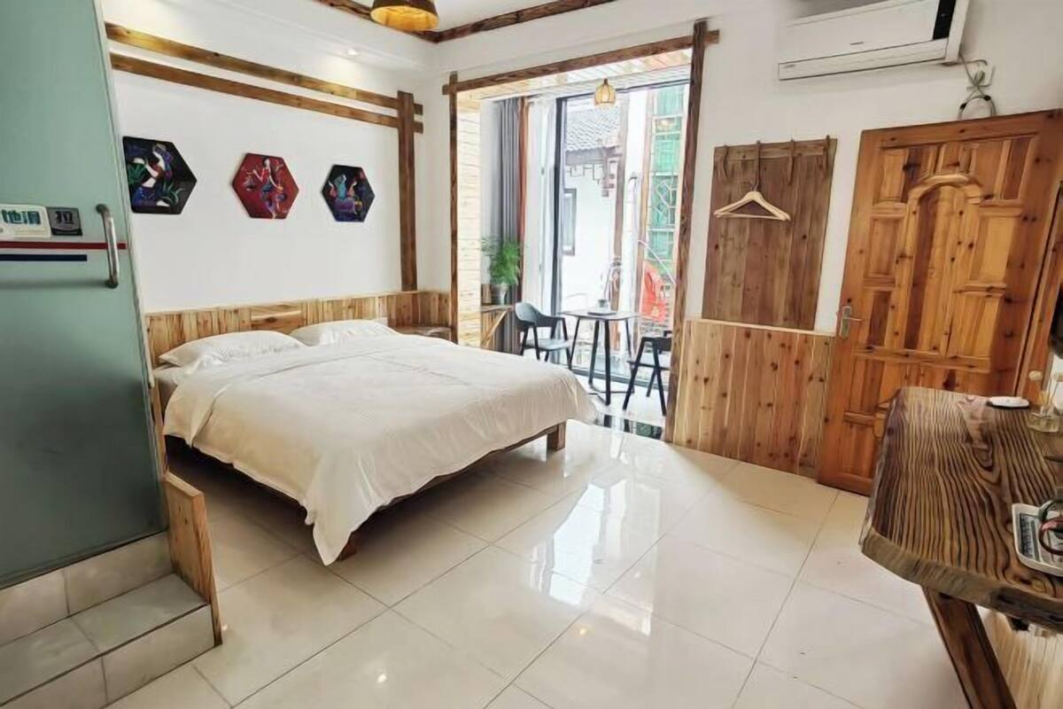 A hotel room with a bed, a wooden wardrobe, a table, and a view of the outside.