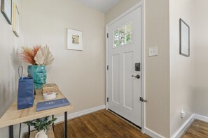 Un couloir avec un plancher en bois, une porte blanche dotée d’une fenêtre, et une table sur laquelle se trouvent un vase, un sac bleu et une boîte.