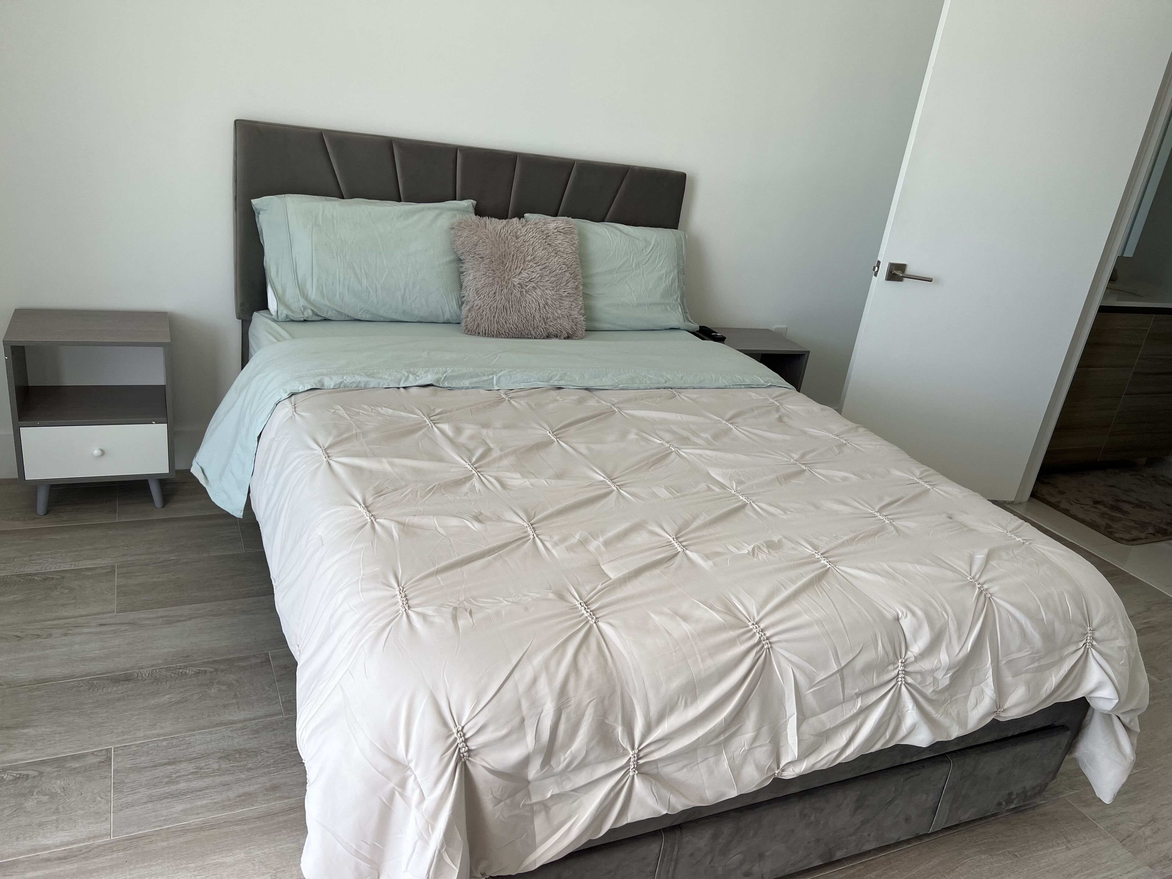 Una cama bien hecha con un edredón blanco capitonado, un cabecero gris y una mesita de noche pequeña con un cajón.