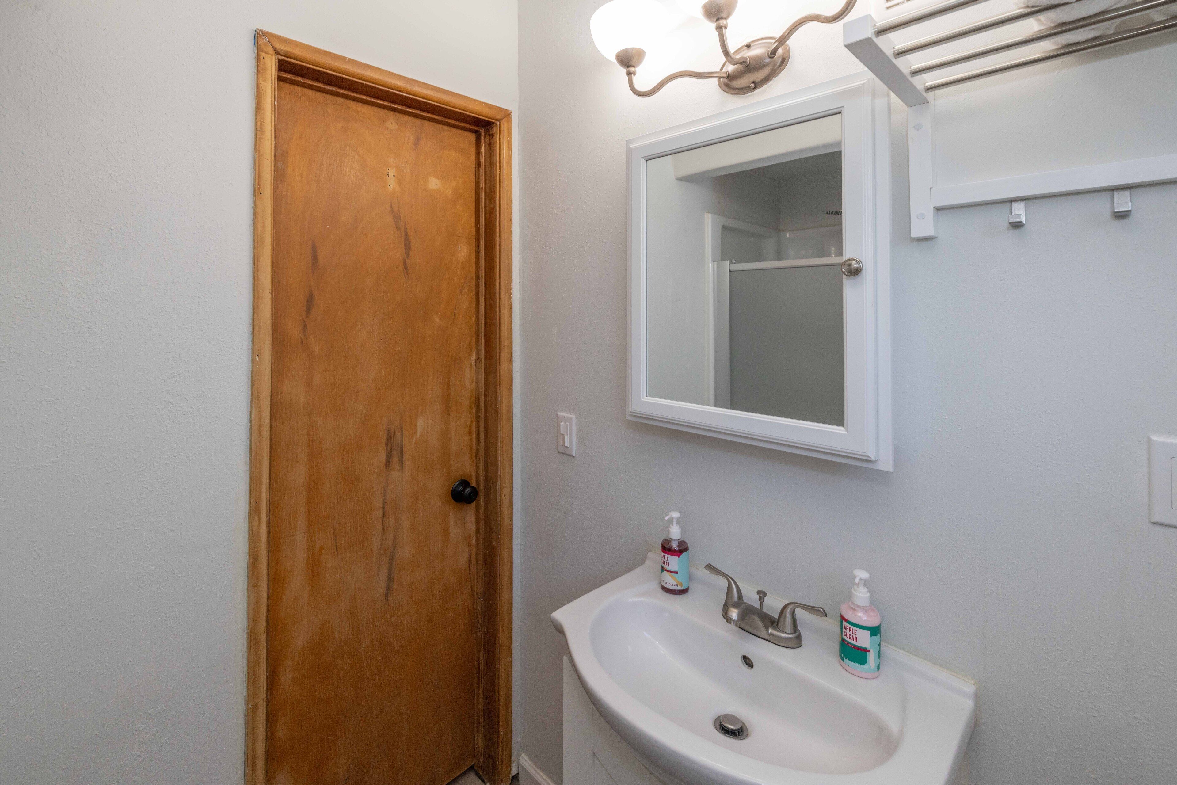 Un baño con puerta de madera, lavabo blanco, espejo y toallero.