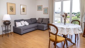 En stue med en grå sofa, et rundt træbord med hvid dug, en lampe og billeder i rammer på væggen.