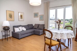 Een woonkamer met een grijze bank, een ronde houten eettafel met een wit tafelkleed, een lamp en ingelijste foto's aan de muur.