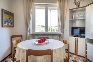 Ein Essbereich mit einem runden Tisch, Stühlen, einer Ablage mit roten Tassen, einem Fenster mit Vorhängen, einem Heizkörper, einem Fernseher auf einem Schrank und einem Gemälde an der Wand.