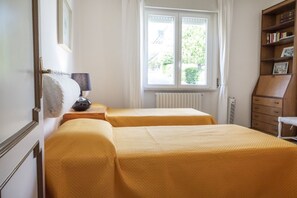 Une chambre avec deux lits, un radiateur, une fenêtre avec des rideaux, une lampe et une bibliothèque.
