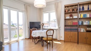 En rummelig stue med spisebord, stole, fjernsyn, reol og store vinduer med gardiner.