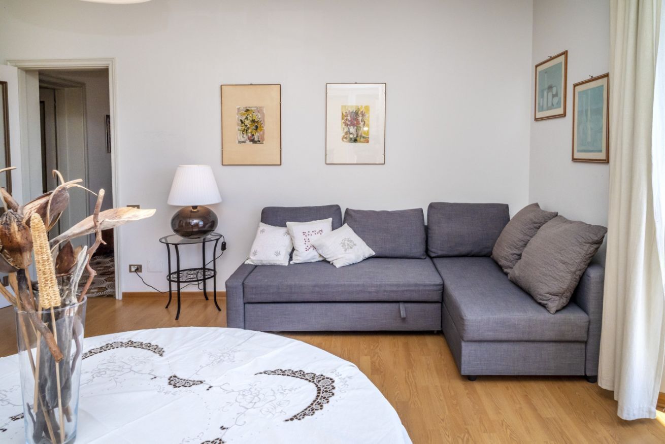 A living room with a grey sofa, a round table with a white tablecloth, a lamp, and framed artwork on the wall.