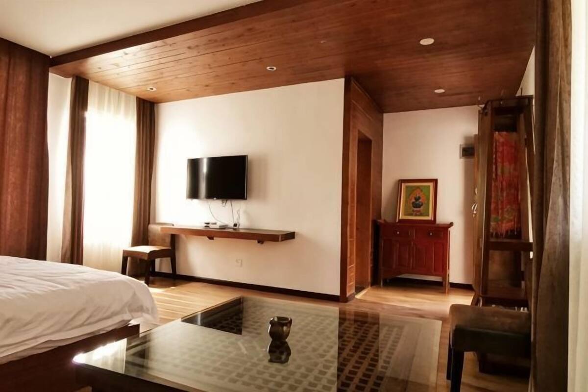 Habitación de hotel con cama, televisor montado en la pared, techo de madera y una pequeña mesa con un jarrón.