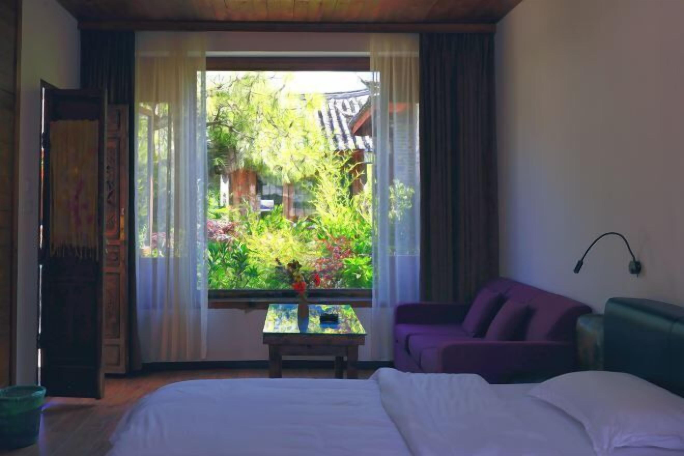 A bedroom with a bed, a purple sofa, a small table with a vase, and a window with a view of greenery and a traditional building.
