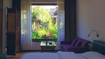 A bedroom with a bed, a purple sofa, a small table with a vase, and a window with a view of greenery and a traditional building.
