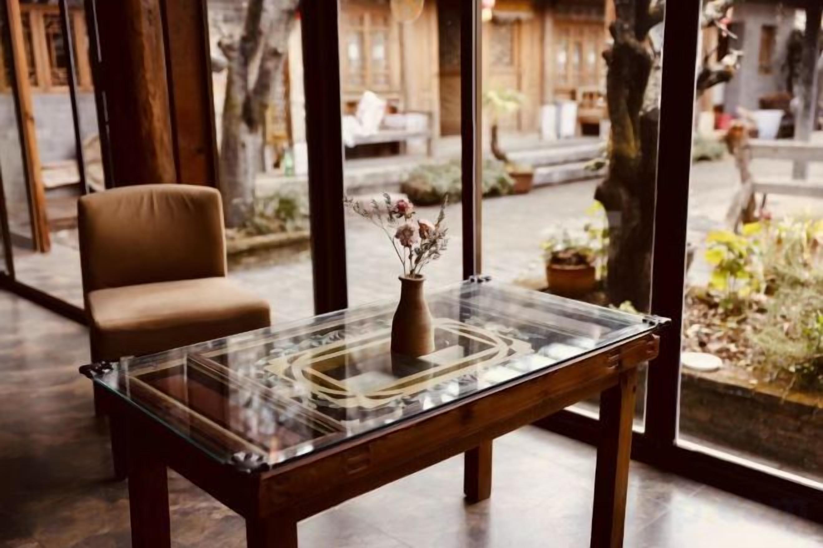 A room with a glass-top table, a vase with flowers, a chair, and a view of the outdoors.